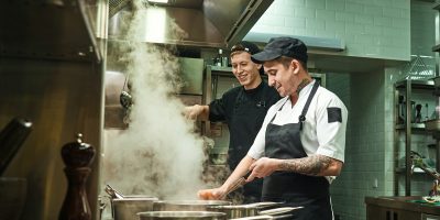 Positive mood. Side view of two cheerful and smiling cooks in uniform are preparing a food in a